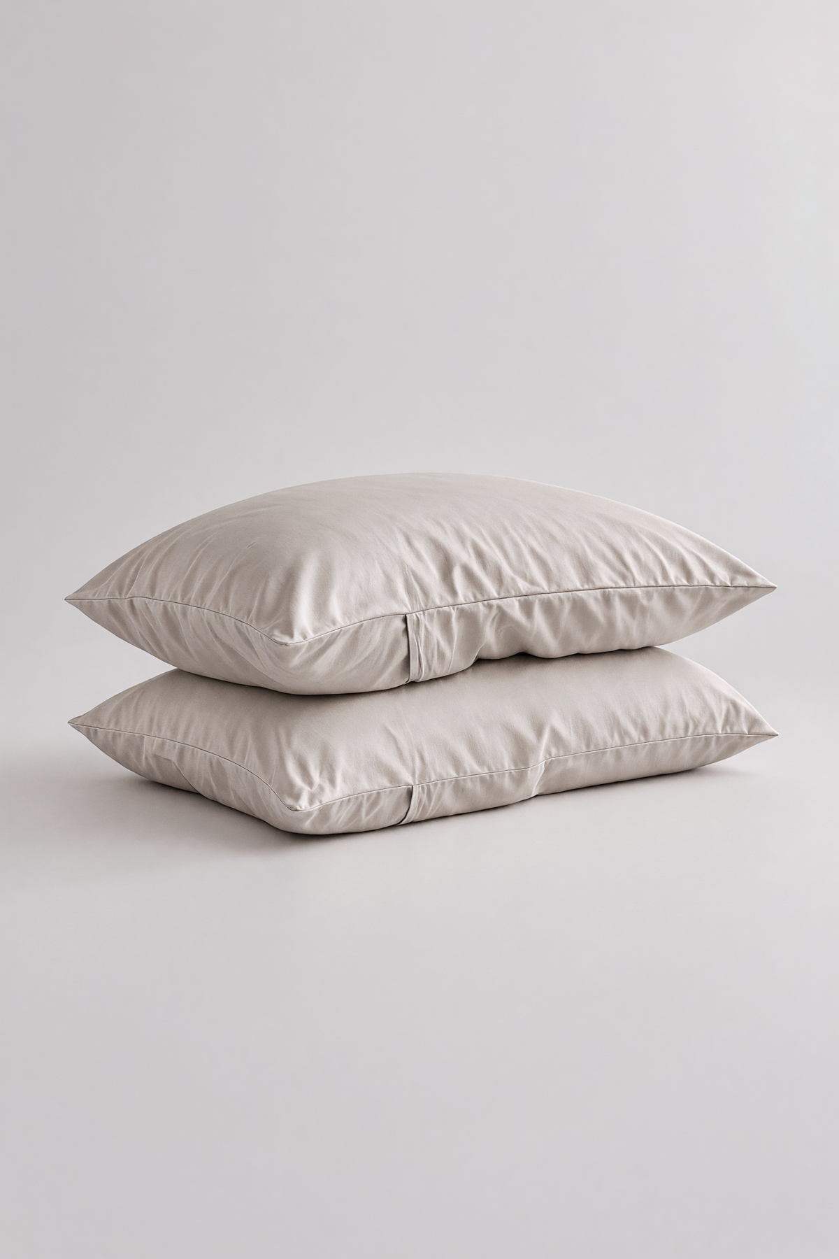 Two plain rectangular pillows stacked on top of each other in a light warm gray or off-white color, photographed against a seamless light gray studio background.