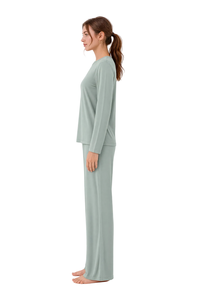 A woman stands in profile wearing a matching two-piece lounge set consisting of a long-sleeve top and wide-leg pants in a soft sage green color, set against a plain white studio background.