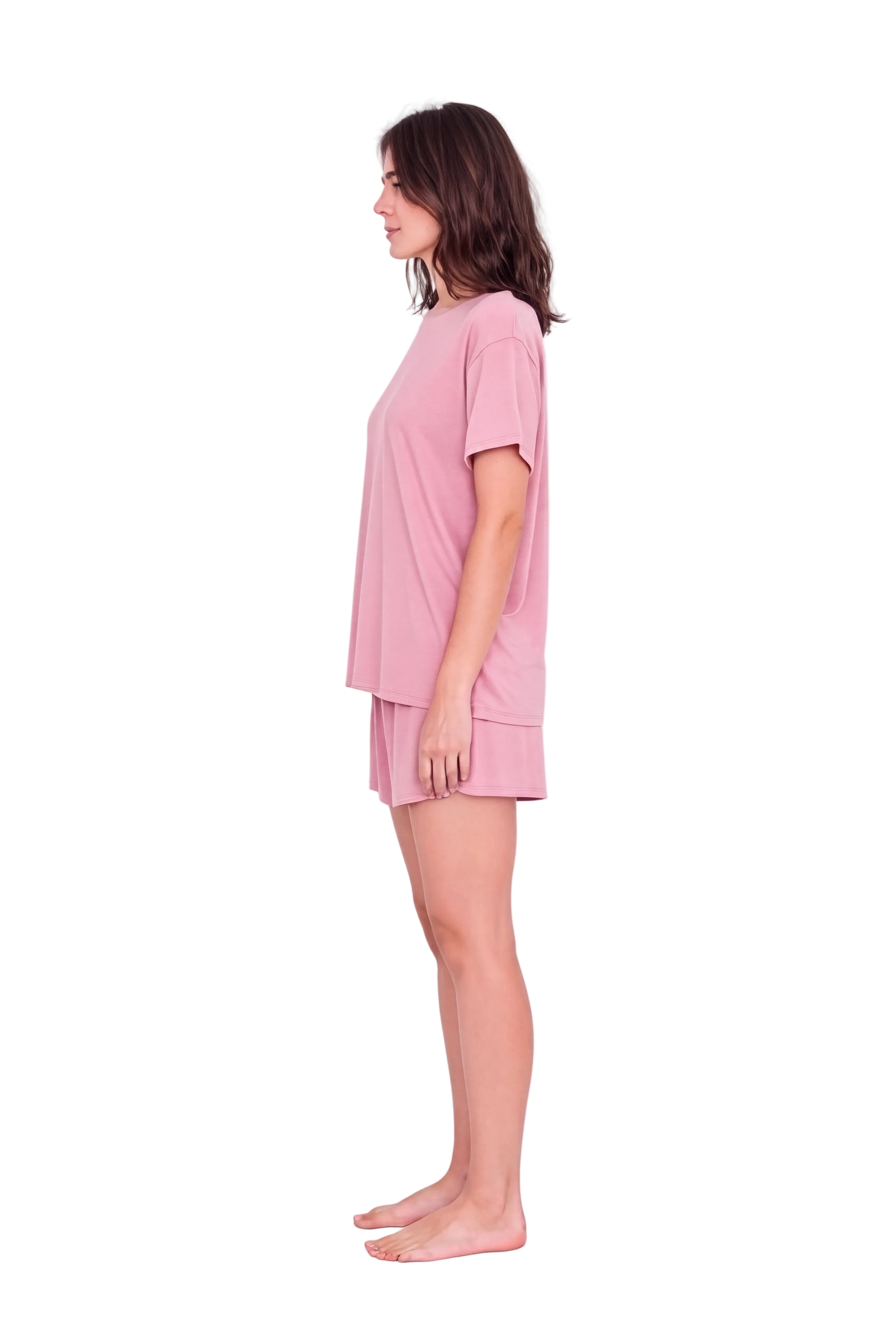 A woman stands in profile wearing a light pink short-sleeve pajama top and matching shorts against a plain light background.
